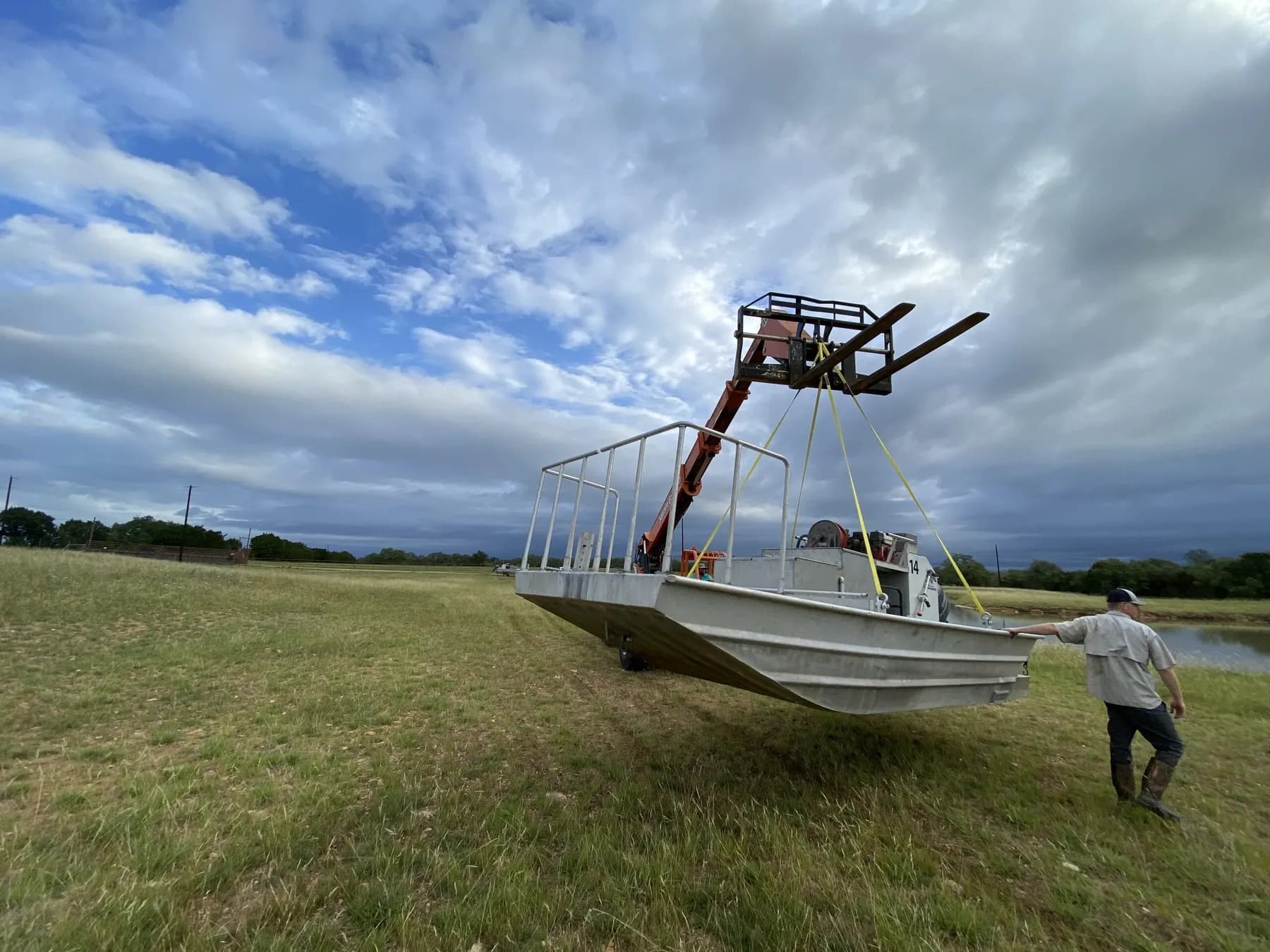 A telehandler crane lifts an electrofishing shock boat by yellow straps across a pasture and over the bank of a lined pond under a stormy Texas sky, while a worker steadies the bow